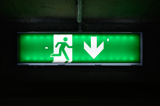 Focus at green electric emergency exit sign on ceiling with blurred background of many people inside of shopping mall.