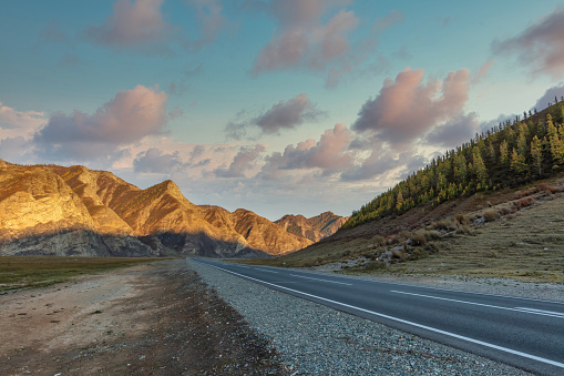 Scenic low angle view of mountain ridge. Highway in the foreground leading all the way up to the mountains. Beautiful cloudy sunset sky as a backdrop. Golden hour. Altai mountains, Siberia, Russia.
