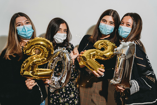 Portrait of four friends celebrating 2021 New Year's Eve together. They are looking at camera and smiling. They are wearing protective face masks.