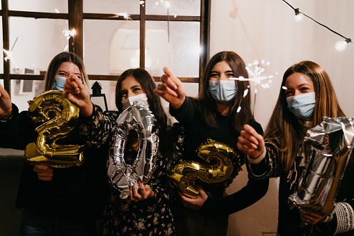 Portrait of four friends celebrating 2021 New Year's Eve together. They are looking at camera, holding sparklers. They are wearing protective face masks.