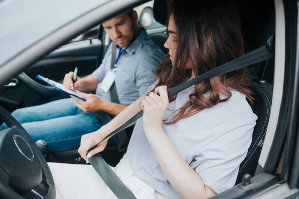 istock Serious young woman fastens her belt. Concentrated driving instructor makes notes on his clipboard. Beautiful young woman with wavy brown hair in white clothes takes driving lessons. 1282510778