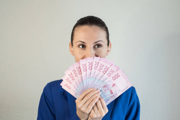 successful businesswoman in suit holding bunch of money banknotes Young woman in classic blue jacket holding banknotes near face isolated on grey background.Portrait of a successful businesswoman in suit holding bunch of money banknotes and looking at camera ukrainian currency stock pictures, royalty-free photos & images