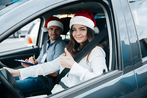 Young woman showing thumb up. Merry Christmas and Happy New Year! Driving courses on winter holidays. Female student and instructor in santa hats smiling and looking into camera through car window