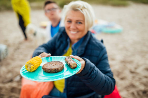 Would You Like A Burger A point of view of a mature woman on her staycation holding up a plate of food and offering something off her plate. family bbq beach stock pictures, royalty-free photos & images