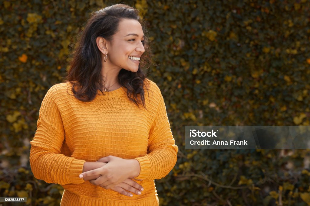 Portrait of beautiful happy woman in a yellow jumper Portrait of beautiful happy woman standing infront of leafy wall with hands on stomach on a lovely autumn day Women Stock Photo