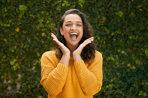 Portrait of beautiful happy woman wearing a yellow jumper and holding up her hands standing infront of green leafy wall