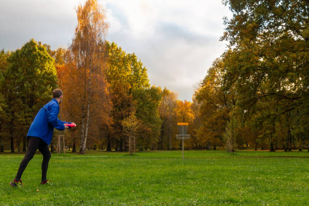 Young caucasian man in blue jacket playing disc golf on autumn play course with basket stock photo