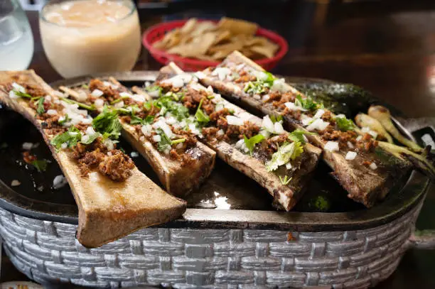 A pewter serving dish arranged with slow-cooked beef bones split to reveal the delicious marrow seasoned with onion, chiles, cilantro,  served with hot fresh corn tortillas at a restaurant in Juarez
