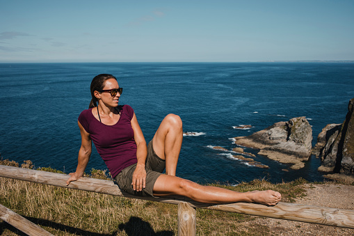 Athletic woman sitting on a wooden railing at seashore in the north of Spain, with Atlantic Ocean in the background
