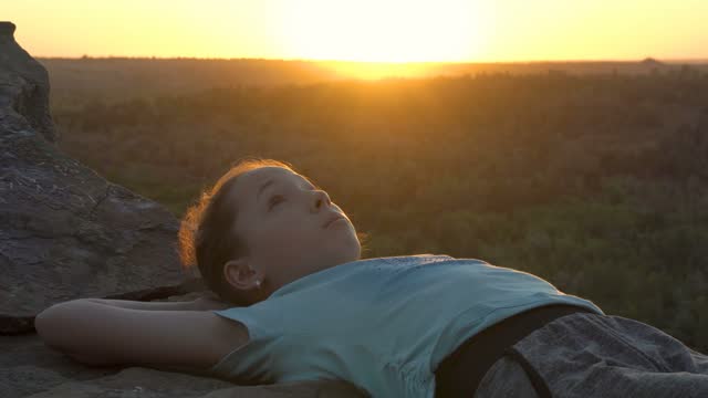 A young teenage girl is lying, looking at the sky and dreaming on a mountain top with a sunset background. Female portrait. Close-up. 4K.