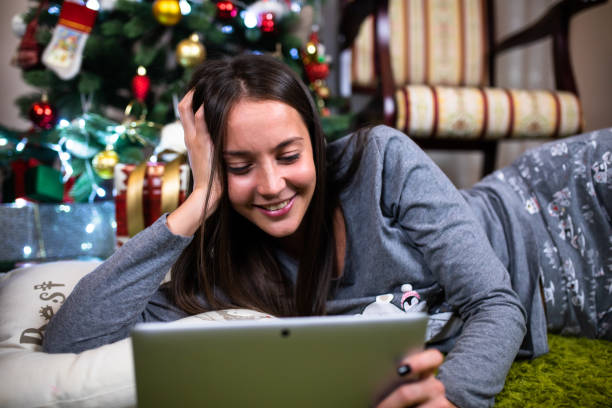 A beautiful woman lying under a Christmas tree looking at a tablet