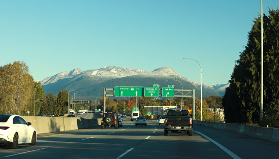Vancouver, Canada - October 24, 2020: Traffic heads north on the Trans Canada Highway through East Vancouver on an autumn morning. Green destination signs point the way to Whistler. Background shows the snow-capped North Shore Mountains.