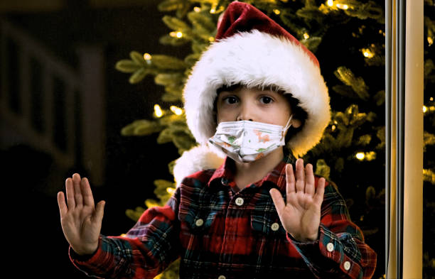 istock Lonely Serious pensive Little boy wearing a Santa hat and a protective face mask looking Through a window at night 1282540096