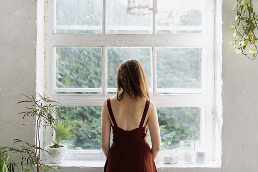 Beautiful adult woman standing near large window, looking at view, spending time in white room with interior design in loft style. Female resting at home, enjoying early morning at weekend