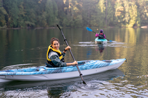 A gorgeous mixed race young adult woman of Pacific Islander descent is happily kayaking on a calm lake and smiling directly at the camera.