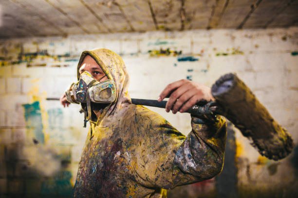 A man with a gas mask is holding a long paint roller in a messy room
