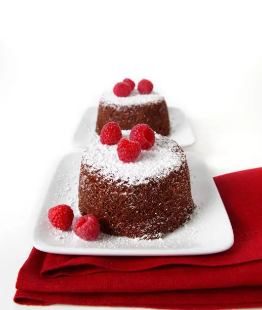 Two individual serving chocolate cakes with raspberries and powdered sugar on top set on a white plate. The plate is top of a red napkin on a white background.