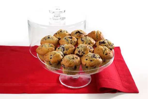 Blueberry muffins on a clear glass cake plate in a red table runner and white background.