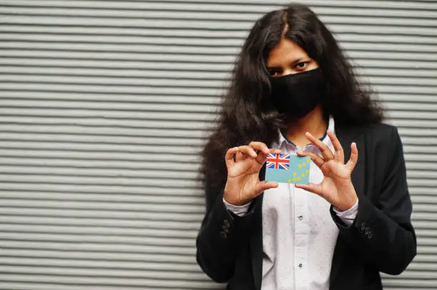 Photo of Asian woman at formal wear and black protect face mask hold Tuvalu flag at hand against gray background. Coronavirus at country concept.