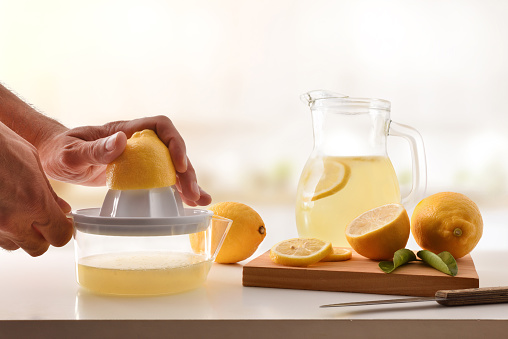 Hands squeezing a lemon in a plastic juicer with container for preparing a lemonade on the kitchen bench. Front view.Horizontal composition.