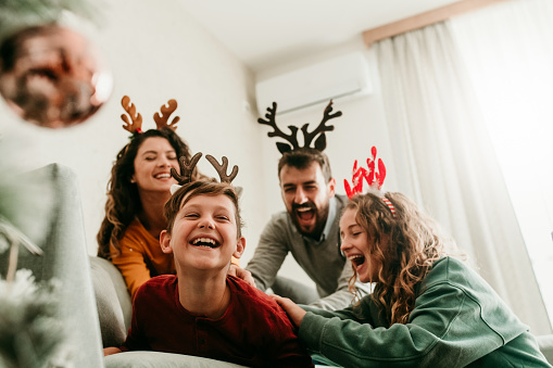 A boy is lying on the sofa in the living room and his family is tickling him on his back. They are laughing and enjoying the time they spend together during the holiday season.