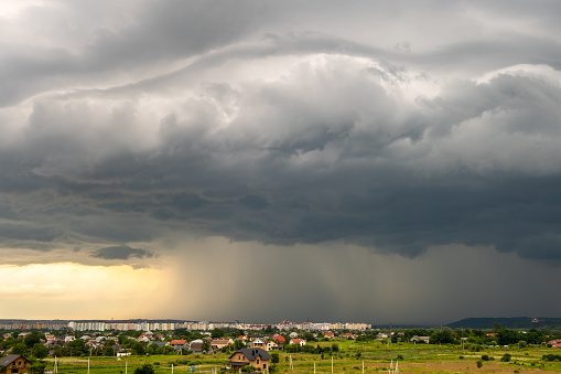 Moody landscape with dark stormy clouds with falling heavy downpour shower rain over distant town buildings in summer.