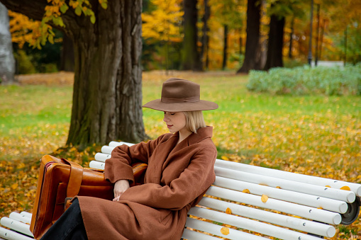 Style girl in brown coat with suitcase sitting on a bench in autumn park