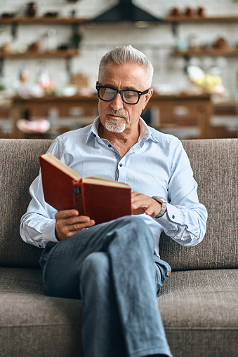 Stylish mature man reading book after hard working day while sitting on cosy sofa. Domestic lifestyle concept