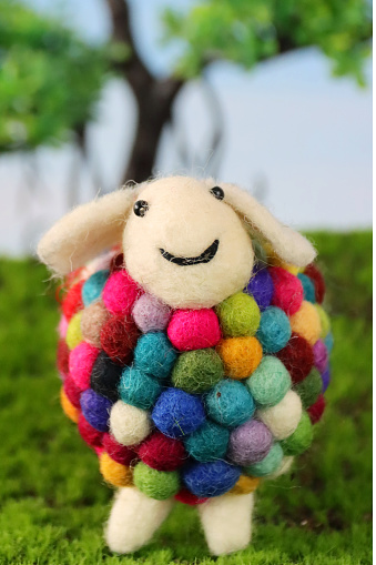 Stock photo showing a close-up view of handmade felt material multicoloured sheep, in front of model tree, in an agricultural farm field scene with green grass and blue sky.