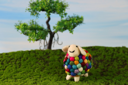 Stock photo showing a close-up view of handmade felt material multicoloured sheep, in front of model tree, in an agricultural farm field scene with green grass and blue sky.