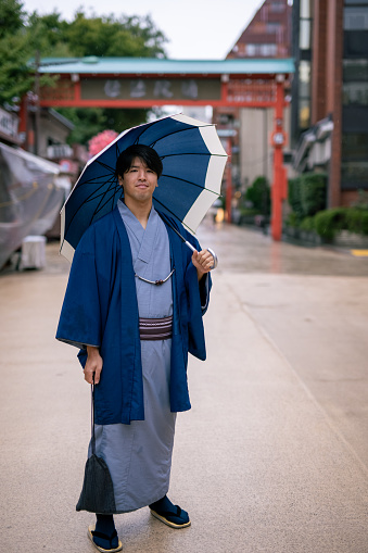 Full length portrait of young man in kimono standing on traditional Japanese street in rainy day