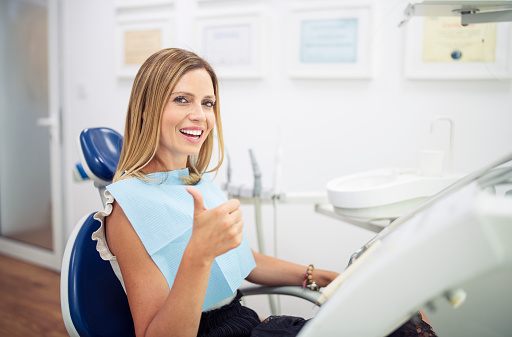 Portrait of beautiful mid adult woman sitting on dentist chair in dental clinic, she smiling and sign thumbs up with hand.