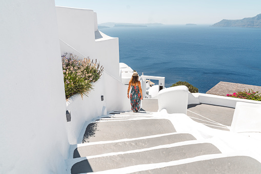 Caucasian woman tourist in long dress with straw hat walking down a staircase in Oia town on a hot summer day. Traditional white houses and streets of Santorini.