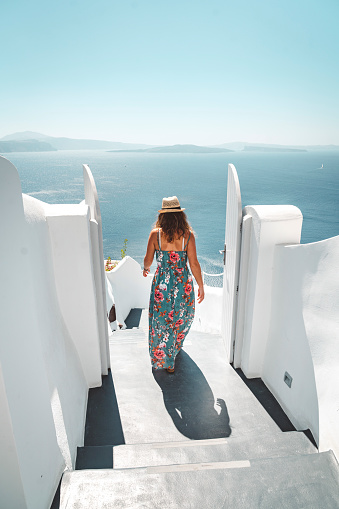 Vertical photo of caucasian woman tourist in long dress with straw hat exploring Santorini island and Oia town on a hot summer day. Traditional white houses and streets of Oia.