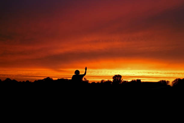 Sunset over fields, distant horizon, trees and buildings Silhouetted against a colorful sky. Person waving goodnight to  the sun.