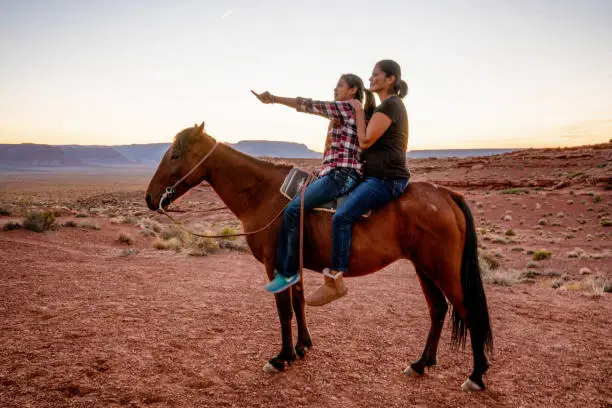 A Navajo Mother And Daughter Riding Their Horse And Looking Over Their Family Land In Monument Valley, Arizona