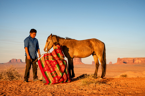 A Native American Young Man Assisting His Beautiful Young Sister With Her Horse, With The Beautiful Monument Valley Behind Them