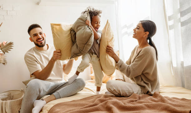 feliz familia multiétnica mamá, papá y niño riendo, jugando, pelea almohadas y saltando en la cama en casa - lucha con almohada fotografías e imágenes de stock