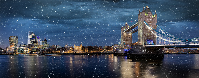 Panoramic view of the illuminated skyline of London, United Kingdom, during winter night time with falling snow over the Tower Bridge and City