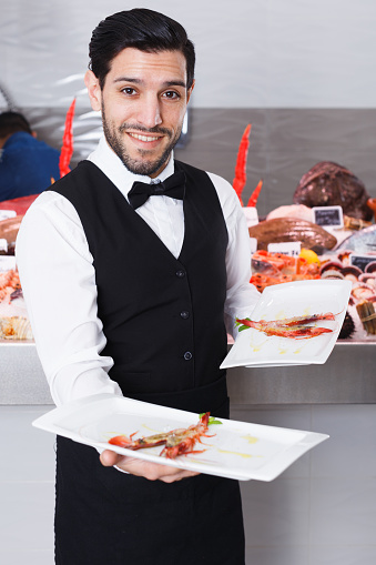 Smiling waiter with seafood dishes in fish restaurant against backdrop of icy showcase