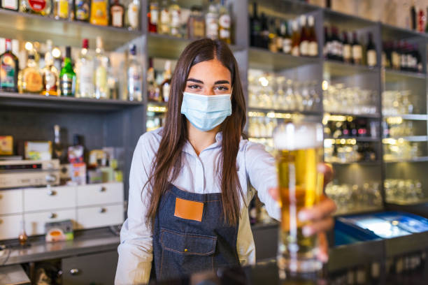 istock Beautiful female Bartender wearing protective face mask, serving a draft beer during coronavirus pandemic, shelves full of bottles with alcohol on the background 1282633709