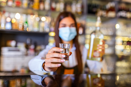 Beautiful female bartender with protective face maskholding shot glass with alcohol during coronavirus pandemic, shelves full of bottles with alcohol on the background