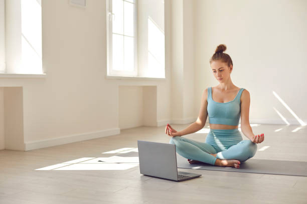 giovane allenatore di yoga seduto a gambe incrociate davanti al computer portatile dando lezione online o webinar di hosting - yoga meeting foto e immagini stock