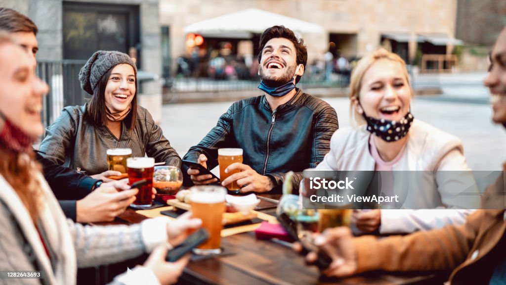 Young friends drinking beer wearing face mask - New normal lifestyle concept with people having fun together talking on happy hour at outside brewery bar - Bright warm filter with focus on central guy Friendship Stock Photo