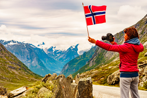 Tourist woman with norwegian flag and camera taking travel picture in mountains on national route Gamle Strynefjellsvegen, Southern Norway. Travel and adventure.