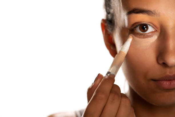 Young dark skinned woman applying concealer on her low eyelids Young dark skinned woman applying concealer on her low eyelids on white background concealer stock pictures, royalty-free photos & images