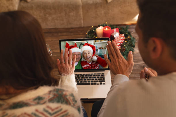 vista posteriore della coppia agitando mentre avendo un videochiamata con coppia senior in cappelli santa sorridente su lapt - santa claus waving christmas photography foto e immagini stock