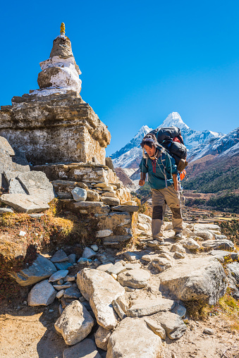 Poerter carrying heavy load around a traditional Buddhist stupa on the Everest Base Camp trail beneath the iconic snowy spire of Ama Dablam (6812m) high in the Himalaya mountain wilderness of the Sagarmatha National Park, a UNESCO World Heritage Site in Nepal.