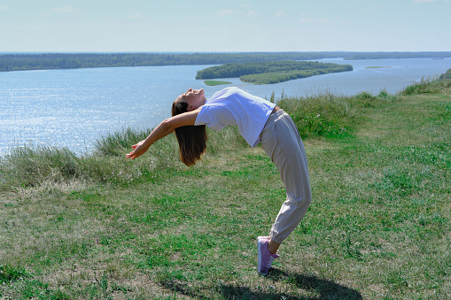 Young sporty woman practicing yoga, standing in Urdhva Dhanurasana exercise, Bridge pose, working out outdoors. happy woman of middle age doing exercises on a river bank alone.