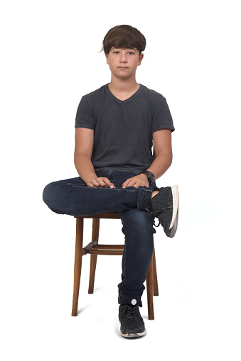 front view of teenage boy sitting on a chair with white background, legs crossed